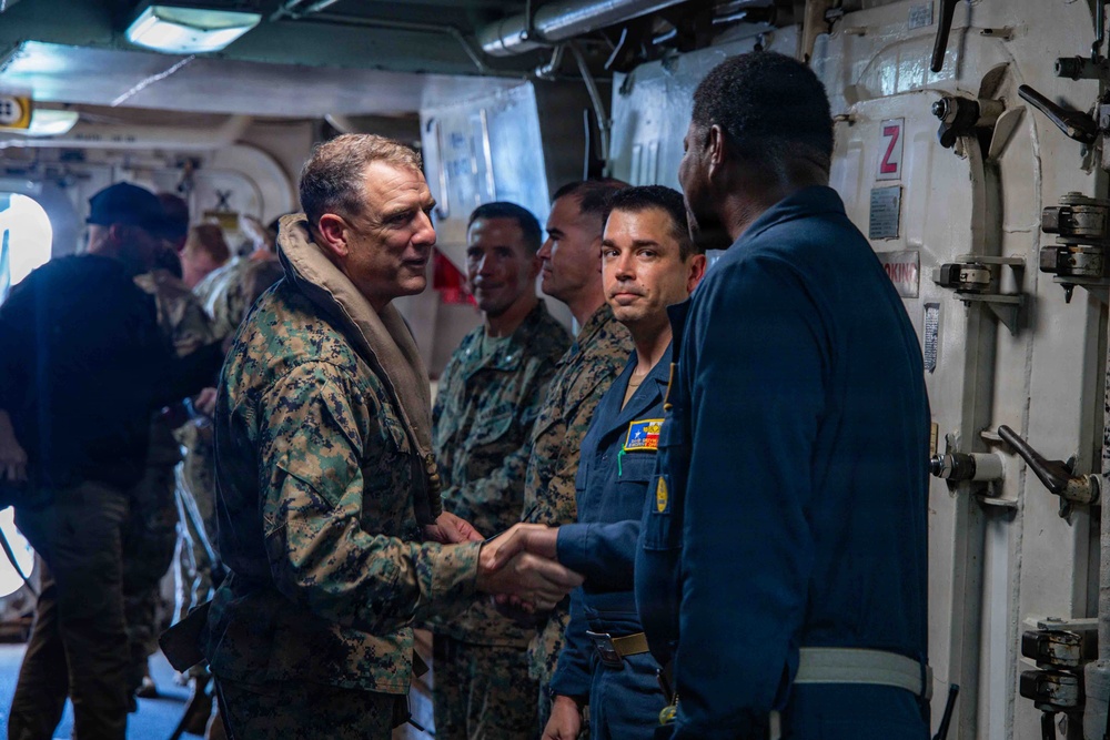 A senior U.S. Marine Corps officer in MARPAT camouflage uniform is shaking hands with a Navy sailor in a blue coverall uniform aboard what appears to be a naval vessel. Several other Marines in camouflage uniforms are present in the background, suggesting an official visit or greeting ceremony. The setting is an interior passageway or compartment of a military ship, identifiable by the watertight door and naval infrastructure visible.