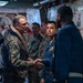 A senior U.S. Marine Corps officer in MARPAT camouflage uniform is shaking hands with a Navy sailor in a blue coverall uniform aboard what appears to be a naval vessel. Several other Marines in camouflage uniforms are present in the background, suggesting an official visit or greeting ceremony. The setting is an interior passageway or compartment of a military ship, identifiable by the watertight door and naval infrastructure visible.