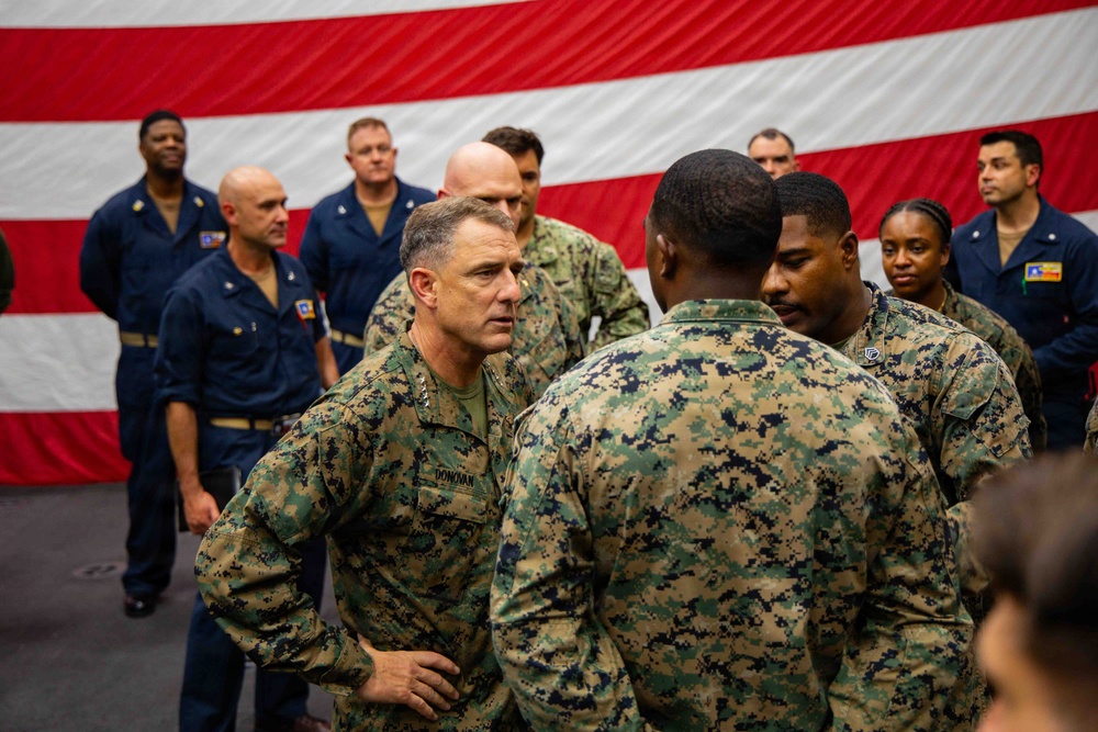 A senior U.S. Marine Corps officer with the name tape 'DONOVAN' and general officer rank insignia is engaged in conversation with two Marines in MARPAT camouflage uniforms. The setting appears to be aboard a naval vessel or military facility, with a large American flag displayed prominently in the background. Navy personnel in blue coveralls are visible standing in the background.