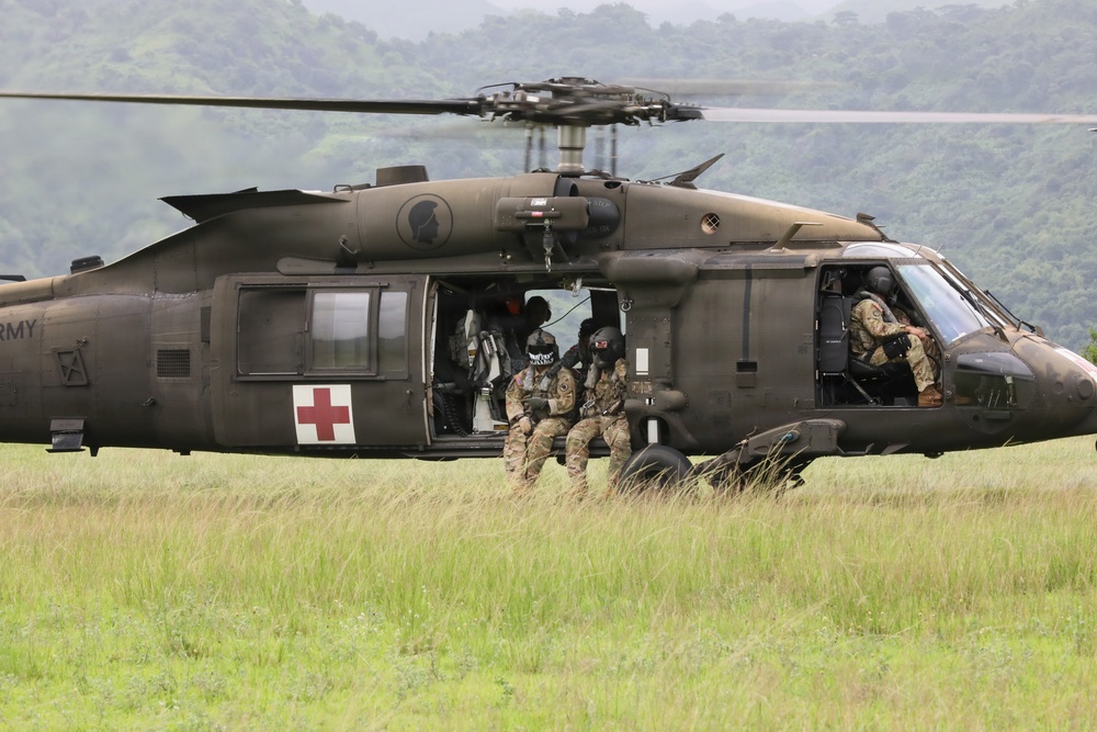 A U.S. Army UH-60 Black Hawk helicopter configured as a MEDEVAC aircraft is landed or hovering low over a grassy field with forested hills in the background. Several soldiers in full combat gear and tactical face coverings are positioned in and around the open side door of the aircraft. The scene appears to depict a medical evacuation exercise or operational insertion/extraction.
