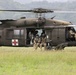 A U.S. Army UH-60 Black Hawk helicopter configured as a MEDEVAC aircraft is landed or hovering low over a grassy field with forested hills in the background. Several soldiers in full combat gear and tactical face coverings are positioned in and around the open side door of the aircraft. The scene appears to depict a medical evacuation exercise or operational insertion/extraction.