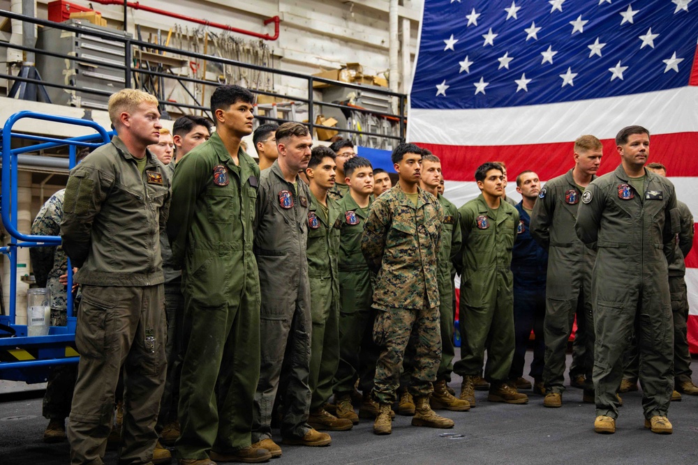 A group of U.S. military personnel, primarily wearing olive green flight suits and one in Marine Corps MARPAT camouflage utilities, are assembled in formation inside what appears to be a ship's hangar bay. A large American flag is prominently displayed in the background. The gathering appears to be a formal ceremony or briefing event.