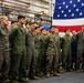 A group of U.S. military personnel, primarily wearing olive green flight suits and one in Marine Corps MARPAT camouflage utilities, are assembled in formation inside what appears to be a ship's hangar bay. A large American flag is prominently displayed in the background. The gathering appears to be a formal ceremony or briefing event.