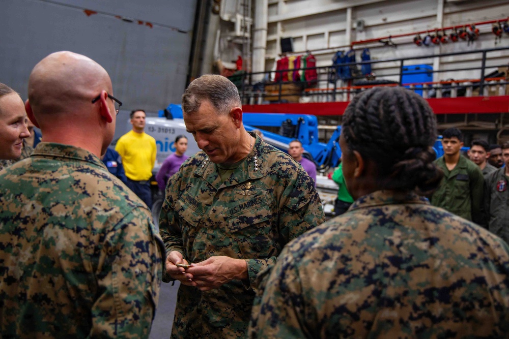 A senior U.S. Marines officer, identifiable by four-star general rank insignia and 'U.S. MARINES' chest tape, appears to be presenting or exchanging a coin with personnel in what looks like a ship's hangar bay. The ceremony involves multiple Marines in MARPAT camouflage uniforms gathered around the general. The setting appears to be aboard a naval vessel, with equipment, scaffolding, and safety gear visible in the background.