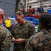 A senior U.S. Marines officer, identifiable by four-star general rank insignia and 'U.S. MARINES' chest tape, appears to be presenting or exchanging a coin with personnel in what looks like a ship's hangar bay. The ceremony involves multiple Marines in MARPAT camouflage uniforms gathered around the general. The setting appears to be aboard a naval vessel, with equipment, scaffolding, and safety gear visible in the background.