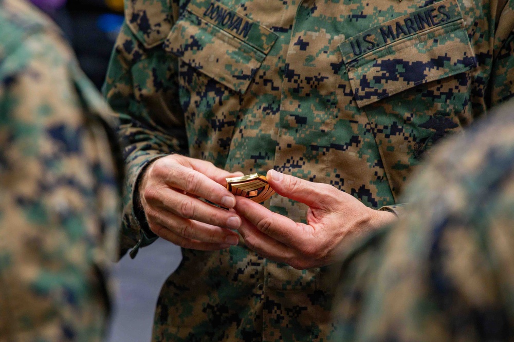 A U.S. Marine wearing MARPAT digital camouflage uniform with the name tape 'DONOVAN' is holding a gold rank insignia device, likely during a promotion or award ceremony. The insignia appears to be a First Sergeant or Sergeant Major chevron device based on its shape and gold color. Another Marine's hands are visible receiving or presenting the insignia.