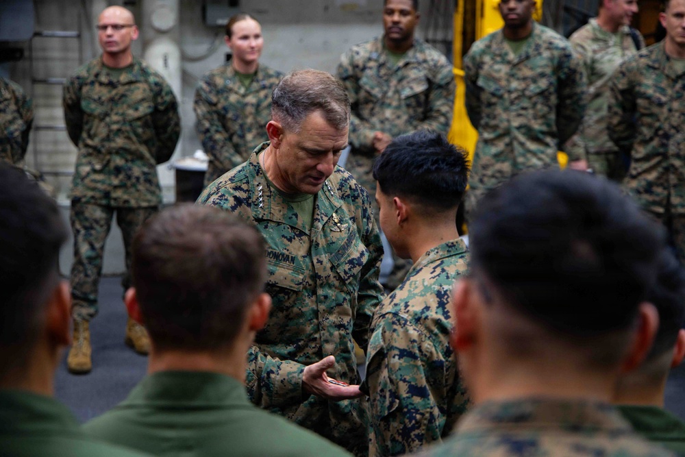 A senior U.S. Marine Corps officer, identified by the name tape 'DONOVAN' and visible general officer rank insignia (stars on collar), appears to be presenting a coin or small item to a junior Marine in what looks like a ship's hangar bay or similar military vessel interior. The ceremony is observed by a group of Marines in MARPAT camouflage uniforms standing in a semicircle. This appears to be a formal coin presentation or recognition ceremony.