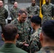 A senior U.S. Marine Corps officer, identified by the name tape 'DONOVAN' and visible general officer rank insignia (stars on collar), appears to be presenting a coin or small item to a junior Marine in what looks like a ship's hangar bay or similar military vessel interior. The ceremony is observed by a group of Marines in MARPAT camouflage uniforms standing in a semicircle. This appears to be a formal coin presentation or recognition ceremony.