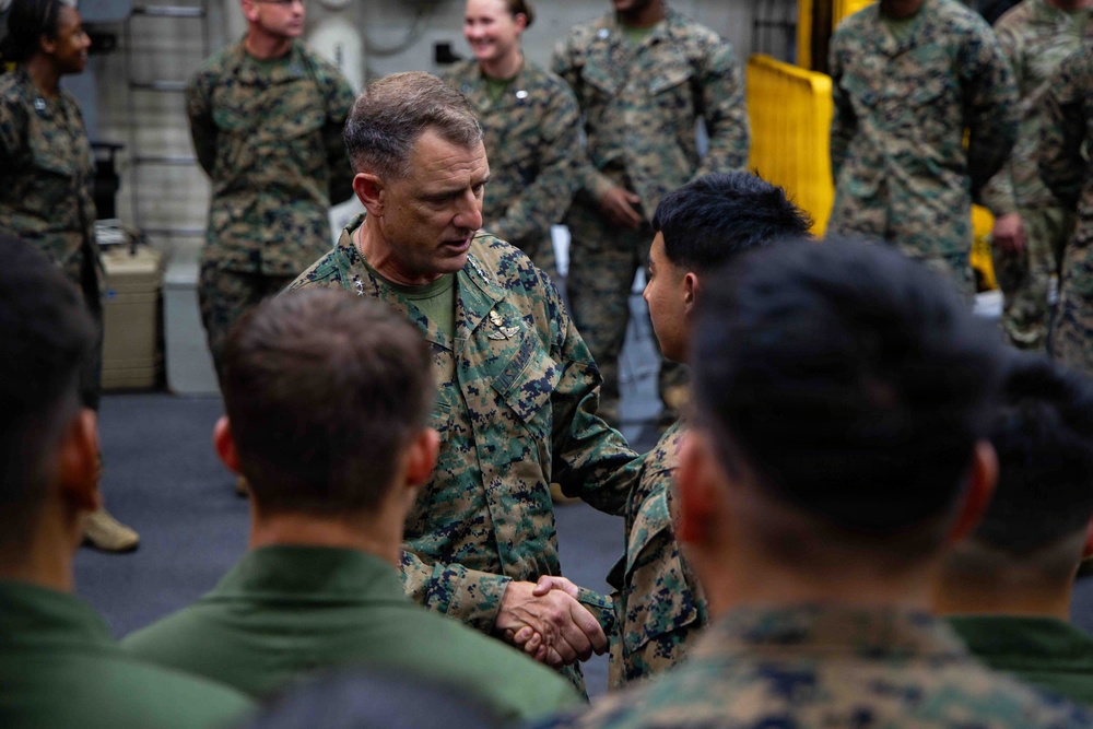 A senior military officer in U.S. Marine Corps MARPAT camouflage uniform is shaking hands with a junior service member, surrounded by a group of Marines in what appears to be a ship's hangar bay or interior deck. The central figure displays rank insignia and a command badge on his uniform, suggesting a formal recognition or greeting ceremony. Multiple Marines are gathered in the background observing the interaction.