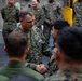 A senior military officer in U.S. Marine Corps MARPAT camouflage uniform is shaking hands with a junior service member, surrounded by a group of Marines in what appears to be a ship's hangar bay or interior deck. The central figure displays rank insignia and a command badge on his uniform, suggesting a formal recognition or greeting ceremony. Multiple Marines are gathered in the background observing the interaction.