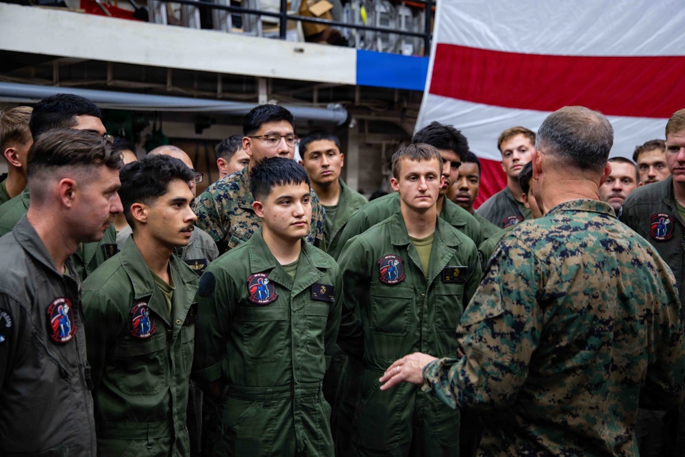 A senior military officer in Marine Corps MARPAT camouflage uniform is addressing a group of service members wearing olive drab flight suits, likely aboard a naval vessel. The gathering appears to be a formal briefing or address, with a large American flag visible in the background. The personnel are attentively listening to the officer who has his back to the camera.
