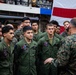 A senior military officer in Marine Corps MARPAT camouflage uniform is addressing a group of service members wearing olive drab flight suits, likely aboard a naval vessel. The gathering appears to be a formal briefing or address, with a large American flag visible in the background. The personnel are attentively listening to the officer who has his back to the camera.