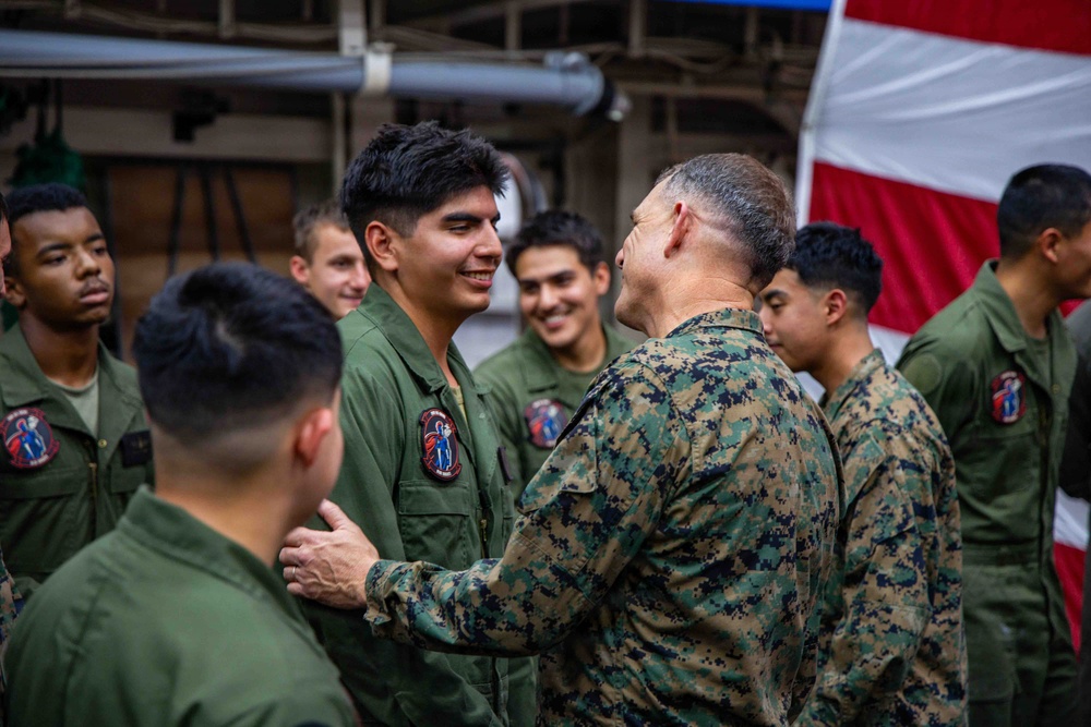 A senior military officer in MARPAT camouflage uniform is interacting with a group of younger service members wearing olive drab flight suits. The setting appears to be inside a military vessel or hangar bay, with a large American flag visible in the background. The interaction appears cordial and positive, possibly a commendation, greeting, or morale visit.