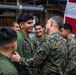 A senior military officer in MARPAT camouflage uniform is interacting with a group of younger service members wearing olive drab flight suits. The setting appears to be inside a military vessel or hangar bay, with a large American flag visible in the background. The interaction appears cordial and positive, possibly a commendation, greeting, or morale visit.
