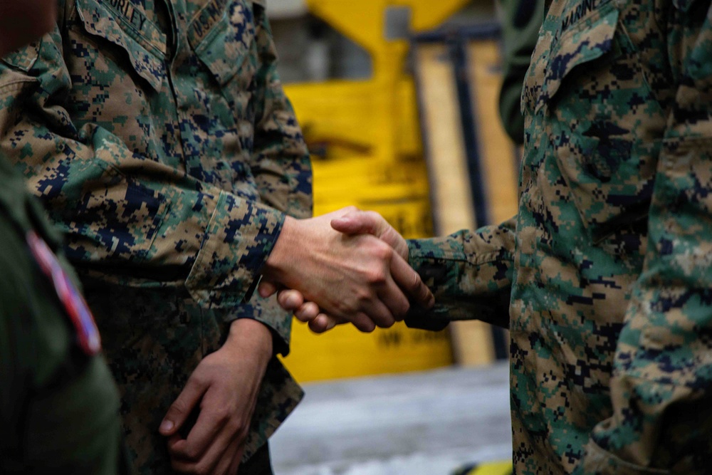 Two individuals wearing U.S. Marine Corps MARPAT (Marine Pattern) digital camouflage uniforms are shaking hands in what appears to be an indoor military facility. The focus is tightly cropped on the handshake, with a yellow piece of equipment or machinery visible in the blurred background. A third person in uniform is partially visible on the left edge of the frame.