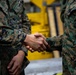 Two individuals wearing U.S. Marine Corps MARPAT (Marine Pattern) digital camouflage uniforms are shaking hands in what appears to be an indoor military facility. The focus is tightly cropped on the handshake, with a yellow piece of equipment or machinery visible in the blurred background. A third person in uniform is partially visible on the left edge of the frame.