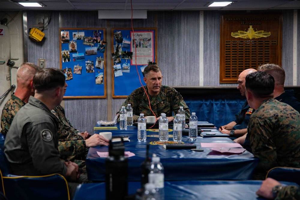 A group of military personnel, primarily in U.S. Marine Corps MARPAT camouflage uniforms, are seated around a conference table in what appears to be a ship's wardroom. A central figure appears to be briefing or leading a discussion. The setting is aboard a U.S. Navy vessel, as indicated by the wardroom placard visible on the wall in the background.
