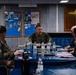 A group of military personnel, primarily in U.S. Marine Corps MARPAT camouflage uniforms, are seated around a conference table in what appears to be a ship's wardroom. A central figure appears to be briefing or leading a discussion. The setting is aboard a U.S. Navy vessel, as indicated by the wardroom placard visible on the wall in the background.