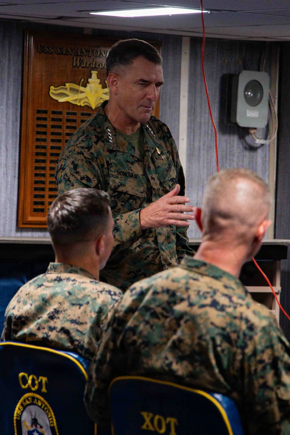 A senior U.S. Marine Corps officer with the name tape 'DONOVAN' and three-star rank insignia (Lieutenant General) is addressing personnel in the wardroom of the USS San Antonio. Two Marines seated with their backs to the camera are listening to the briefing. The setting is an official military meeting aboard a U.S. Navy amphibious transport dock ship.