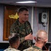 A senior U.S. Marine Corps officer with the name tape 'DONOVAN' and three-star rank insignia (Lieutenant General) is addressing personnel in the wardroom of the USS San Antonio. Two Marines seated with their backs to the camera are listening to the briefing. The setting is an official military meeting aboard a U.S. Navy amphibious transport dock ship.