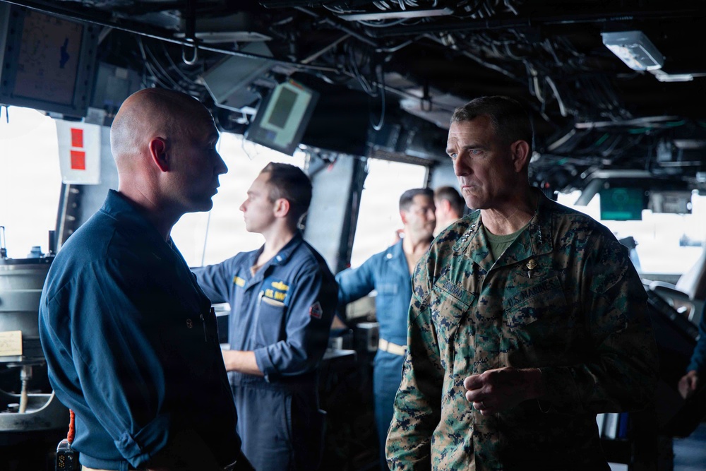 The image shows military personnel aboard what appears to be a naval vessel's bridge or command center. A U.S. Marine Corps officer in MARPAT camouflage uniform is engaged in conversation with a Navy officer in a dark blue uniform. Several other Navy personnel are visible in the background operating equipment.