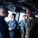 The image shows military personnel aboard what appears to be a naval vessel's bridge or command center. A U.S. Marine Corps officer in MARPAT camouflage uniform is engaged in conversation with a Navy officer in a dark blue uniform. Several other Navy personnel are visible in the background operating equipment.