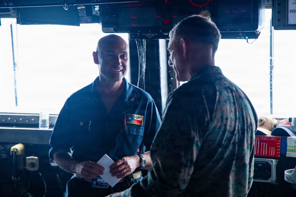 Two military personnel are engaged in conversation aboard what appears to be a naval vessel's bridge or command area. One individual is wearing a dark blue Navy coverall uniform with a name tag and patch, while the other is wearing Marine Corps MARPAT camouflage utilities. The setting includes various navigation and electronic equipment consistent with a ship's bridge.