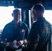 Two military personnel are engaged in conversation aboard what appears to be a naval vessel's bridge or command area. One individual is wearing a dark blue Navy coverall uniform with a name tag and patch, while the other is wearing Marine Corps MARPAT camouflage utilities. The setting includes various navigation and electronic equipment consistent with a ship's bridge.