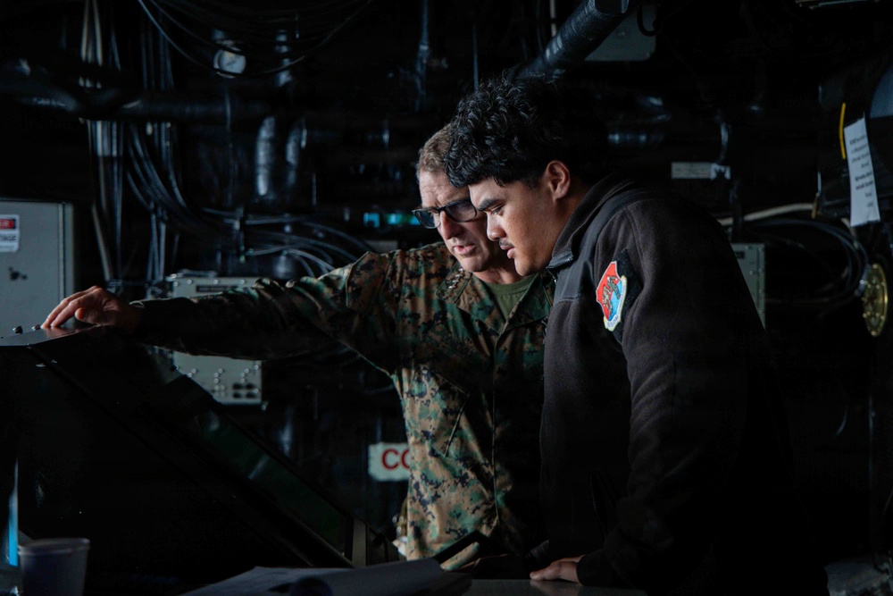 Two individuals are examining equipment in what appears to be a military or naval technical space. One person is wearing U.S. Marine Corps camouflage (MARPAT) uniform with glasses, while the other wears a black jacket with a military unit patch. They appear to be conducting a review or inspection of technical equipment in a dimly lit compartment.