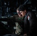 Two individuals are examining equipment in what appears to be a military or naval technical space. One person is wearing U.S. Marine Corps camouflage (MARPAT) uniform with glasses, while the other wears a black jacket with a military unit patch. They appear to be conducting a review or inspection of technical equipment in a dimly lit compartment.