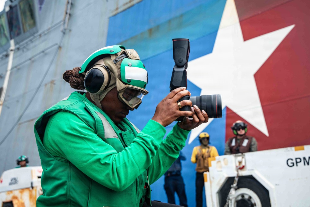 A naval personnel member wearing a green flight deck jersey and cranial helmet with hearing protection holds a professional DSLR camera with a telephoto lens on what appears to be an aircraft carrier flight deck. In the background, a large vessel displays a prominent red, white, and blue star emblem on its hull, along with other crew members in various colored jerseys. The setting indicates a military maritime operation, likely a replenishment-at-sea or port visit.