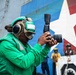 A naval personnel member wearing a green flight deck jersey and cranial helmet with hearing protection holds a professional DSLR camera with a telephoto lens on what appears to be an aircraft carrier flight deck. In the background, a large vessel displays a prominent red, white, and blue star emblem on its hull, along with other crew members in various colored jerseys. The setting indicates a military maritime operation, likely a replenishment-at-sea or port visit.