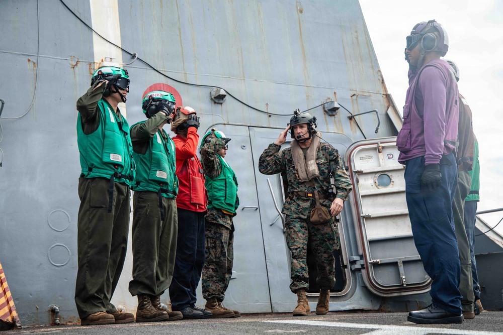 Military personnel are gathered on the deck of a naval vessel, with several individuals wearing flight deck gear including green vests, helmets, and goggles. A Marine in camouflage utilities and a flight helmet appears to be communicating or receiving instructions, while two civilians in casual clothing observe from the right. The scene suggests a flight deck briefing or operational coordination exercise aboard a warship.