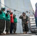 Military personnel are gathered on the deck of a naval vessel, with several individuals wearing flight deck gear including green vests, helmets, and goggles. A Marine in camouflage utilities and a flight helmet appears to be communicating or receiving instructions, while two civilians in casual clothing observe from the right. The scene suggests a flight deck briefing or operational coordination exercise aboard a warship.
