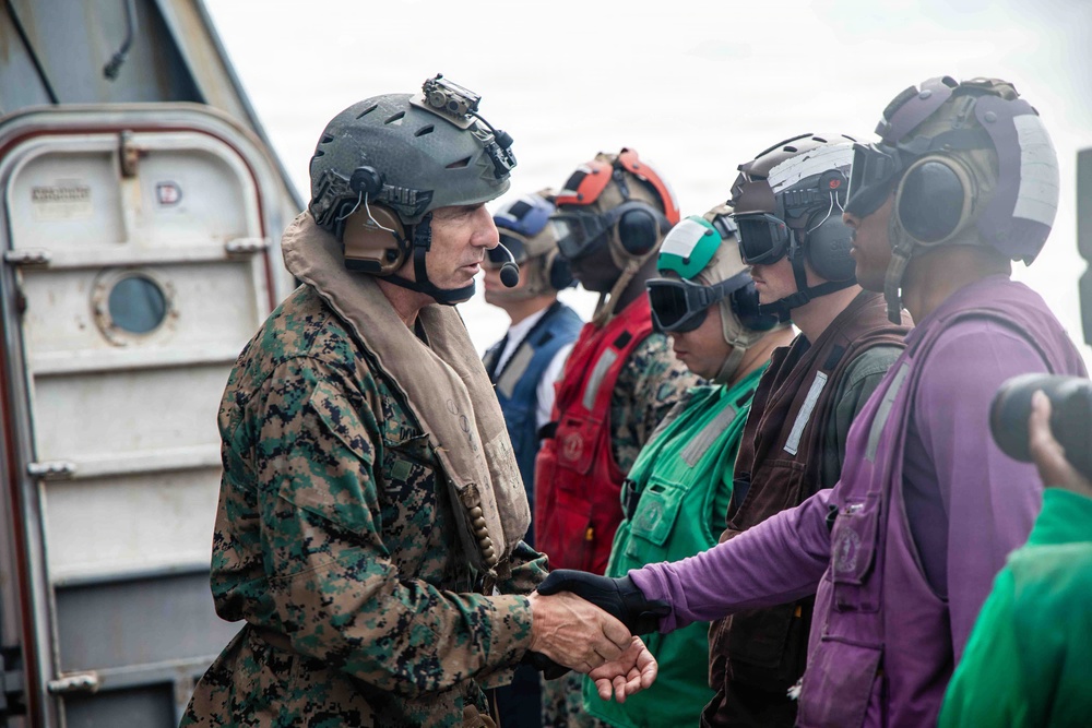 A U.S. Marine in digital camouflage uniform and tactical helmet is shaking hands with personnel wearing colored flight deck jerseys and helmets, likely aboard a naval vessel. The scene appears to depict a greeting or briefing interaction on what appears to be a ship's flight deck or exterior deck area. Multiple personnel in various colored jerseys (green, red, purple) and hearing protection are present, indicating a naval aviation environment.