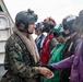 A U.S. Marine in digital camouflage uniform and tactical helmet is shaking hands with personnel wearing colored flight deck jerseys and helmets, likely aboard a naval vessel. The scene appears to depict a greeting or briefing interaction on what appears to be a ship's flight deck or exterior deck area. Multiple personnel in various colored jerseys (green, red, purple) and hearing protection are present, indicating a naval aviation environment.