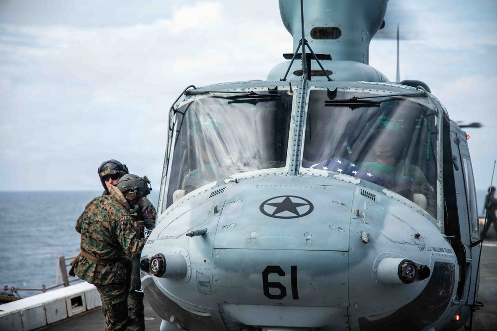 A military helicopter marked with the number '61' and a star insignia is positioned on what appears to be a naval vessel deck over open ocean. A Marine in woodland camouflage uniform and flight helmet is standing beside the helicopter, appearing to communicate with or inspect the aircraft. An American flag is visible through the cockpit windscreen.