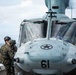 A military helicopter marked with the number '61' and a star insignia is positioned on what appears to be a naval vessel deck over open ocean. A Marine in woodland camouflage uniform and flight helmet is standing beside the helicopter, appearing to communicate with or inspect the aircraft. An American flag is visible through the cockpit windscreen.