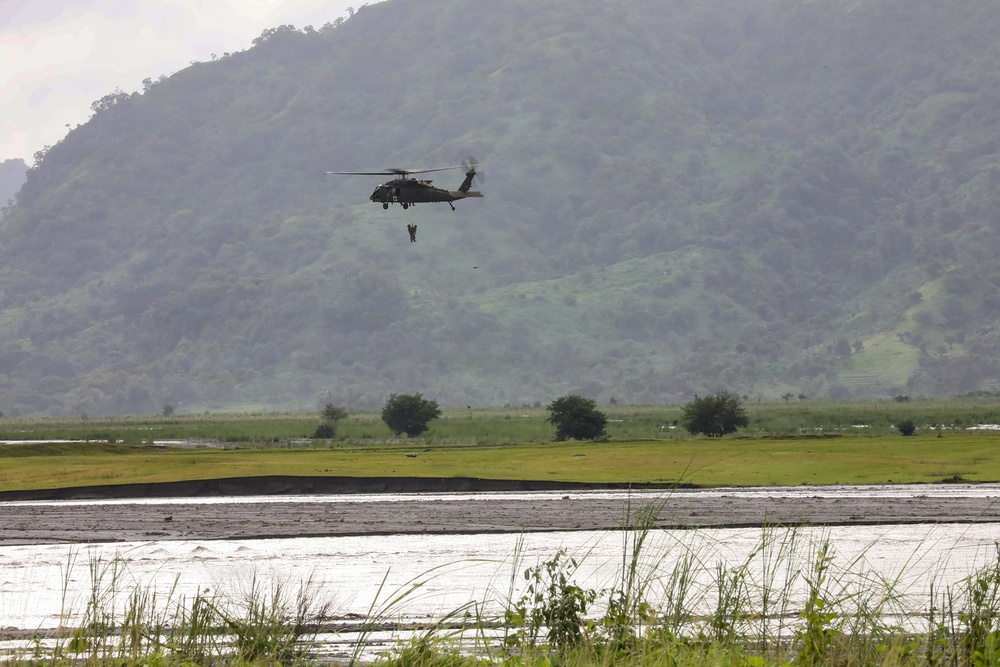 A military helicopter, appearing to be a UH-60 Black Hawk, is hovering over a flooded lowland area near a river with a figure suspended below it on a hoist or rope. The scene suggests a rescue or training operation taking place over waterlogged terrain with a green, forested hillside in the background. The environment shows signs of flooding with muddy, swollen waterways visible in the foreground.