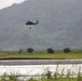 A military helicopter, appearing to be a UH-60 Black Hawk, is hovering over a flooded lowland area near a river with a figure suspended below it on a hoist or rope. The scene suggests a rescue or training operation taking place over waterlogged terrain with a green, forested hillside in the background. The environment shows signs of flooding with muddy, swollen waterways visible in the foreground.