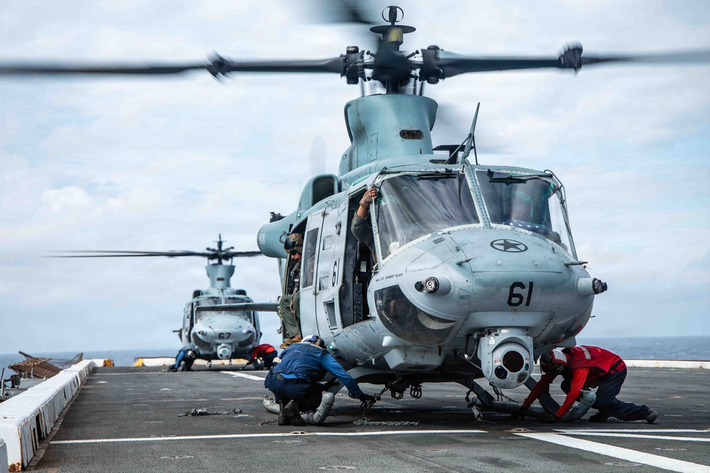 A U.S. Marine Corps UH-1Y Venom helicopter (tail number 61) is on the flight deck of a naval vessel, with rotor blades spinning. Ground crew personnel are working around the aircraft, appearing to secure or release tie-down chains. A second helicopter (tail number 67) is visible in the background on the same flight deck.