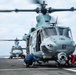 A U.S. Marine Corps UH-1Y Venom helicopter (tail number 61) is on the flight deck of a naval vessel, with rotor blades spinning. Ground crew personnel are working around the aircraft, appearing to secure or release tie-down chains. A second helicopter (tail number 67) is visible in the background on the same flight deck.