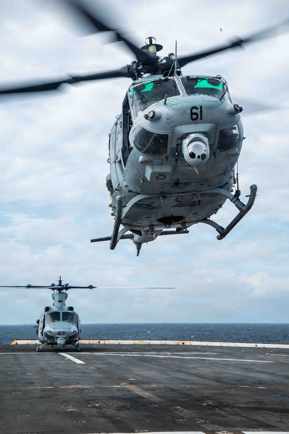 Two military helicopters are operating on the flight deck of a naval vessel at sea. The foreground helicopter, numbered '61', is airborne and hovering low over the deck, while a second helicopter numbered '67' remains on the deck in the background. Both aircraft appear to be UH-1Y Venom helicopters used by the United States Marine Corps.