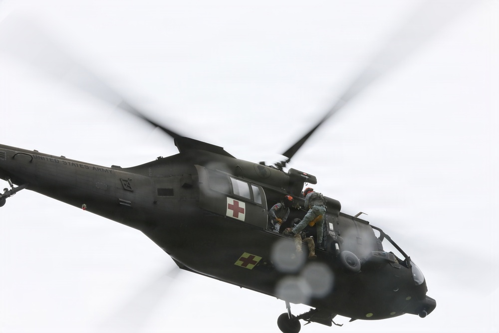 A United States Army medical evacuation (MEDEVAC) helicopter, identifiable as a UH-60 Black Hawk, is captured in flight against an overcast sky. The aircraft bears red cross markings indicating its medical role, and at least one uniformed personnel is visible in the open side door. The image shows motion blur on the rotor blades, indicating active flight.