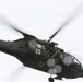 A United States Army medical evacuation (MEDEVAC) helicopter, identifiable as a UH-60 Black Hawk, is captured in flight against an overcast sky. The aircraft bears red cross markings indicating its medical role, and at least one uniformed personnel is visible in the open side door. The image shows motion blur on the rotor blades, indicating active flight.