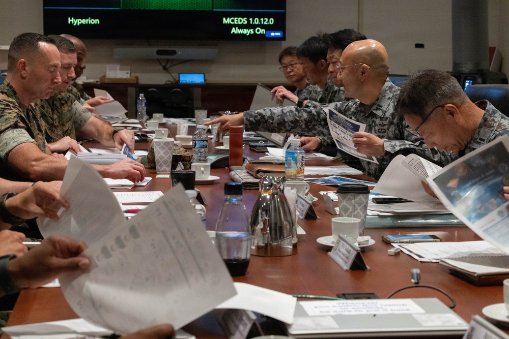 A formal military briefing or staff meeting is taking place at a large conference table, involving personnel in two distinct uniforms — US Marine Corps camouflage (MARPAT) on the left and what appears to be Japan Self-Defense Force uniforms on the right. Participants are reviewing printed documents and briefing materials. A display screen in the background shows 'Hyperion' and 'MCEDS 1.0.12.0 Always On' text.