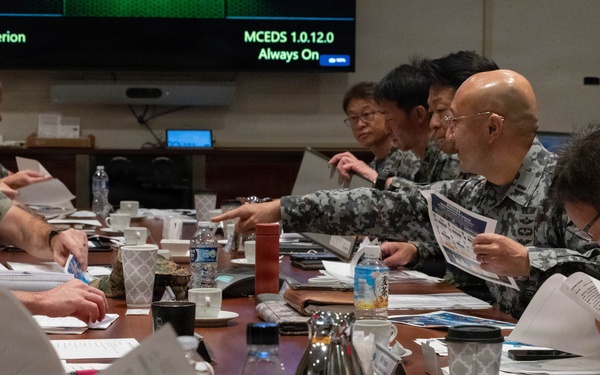 A formal military briefing or staff meeting is taking place at a large conference table, involving personnel in two distinct uniforms — US Marine Corps camouflage (MARPAT) on the left and what appears to be Japan Self-Defense Force uniforms on the right. Participants are reviewing printed documents and briefing materials. A display screen in the background shows 'Hyperion' and 'MCEDS 1.0.12.0 Always On' text.