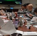 A formal military briefing or staff meeting is taking place at a large conference table, involving personnel in two distinct uniforms — US Marine Corps camouflage (MARPAT) on the left and what appears to be Japan Self-Defense Force uniforms on the right. Participants are reviewing printed documents and briefing materials. A display screen in the background shows 'Hyperion' and 'MCEDS 1.0.12.0 Always On' text.