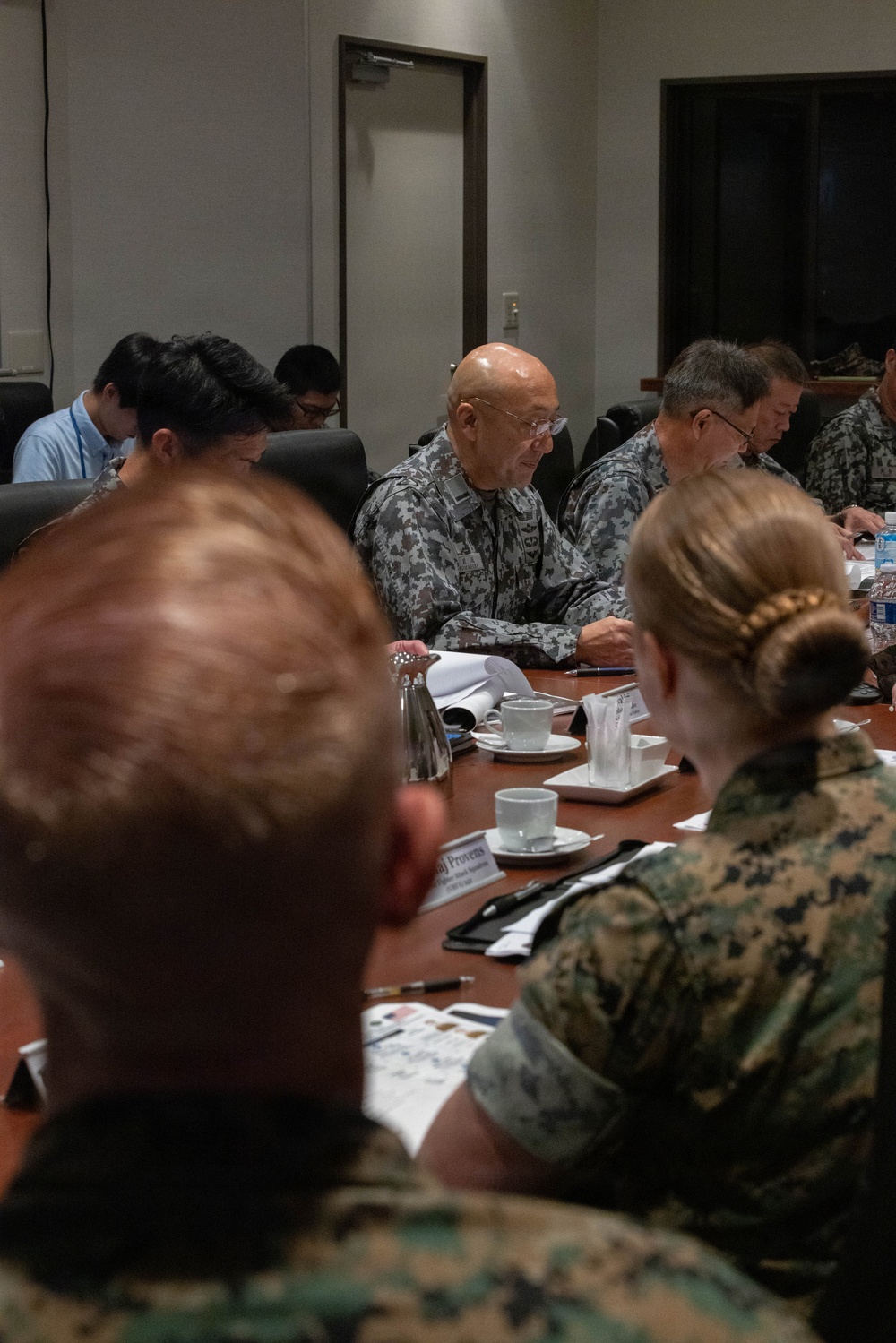 A formal military conference or briefing is taking place around a large wooden conference table. Personnel in multiple camouflage uniform patterns, including what appears to be USMC MARPAT and an Asian military digital camouflage pattern, are seated together alongside civilian attendees. The meeting appears to be a multinational military coordination session with documents, name placards, and beverages on the table.