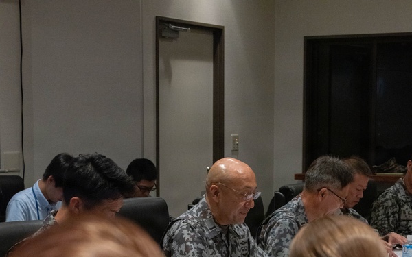 A formal military conference or briefing is taking place around a large wooden conference table. Personnel in multiple camouflage uniform patterns, including what appears to be USMC MARPAT and an Asian military digital camouflage pattern, are seated together alongside civilian attendees. The meeting appears to be a multinational military coordination session with documents, name placards, and beverages on the table.
